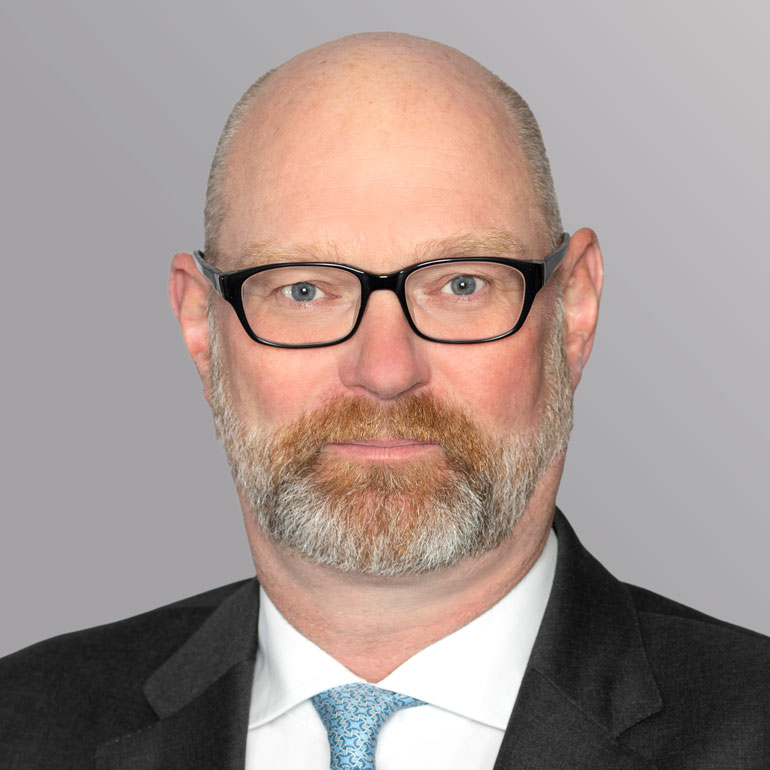 Ein Mann mit einer Glatze und einem gepflegten Bart trägt eine Brille und einen Anzug. Er schaut direkt in die Kamera und hat einen neutralen Gesichtsausdruck. Der Hintergrund ist einfarbig grau.