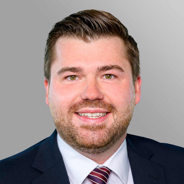 Ein lächelnder Mann mit braunem, kurzem Haar und einem Bart, trägt einen Anzug mit einem hellen Hemd und einer gestreiften Krawatte. Der Hintergrund ist graufarben.