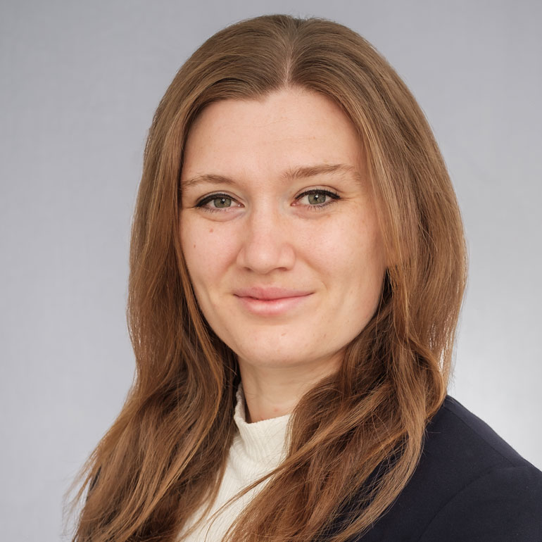 Porträt einer jungen Frau mit langen, brünetten Haaren, die in die Kamera lächelt. Sie trägt einen dunklen Blazer über einem hellen Pullover. Der Hintergrund ist grau und neutral gehalten. Ihre Gesichtszüge sind freundlich und selbstbewusst.