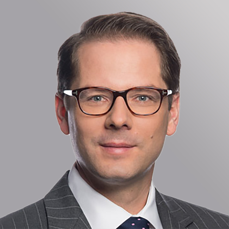 Ein Mann mit kurzen, braunen Haaren und einer Brille lächelt in die Kamera. Er trägt einen grauen Anzug mit feinen Linien und ein blaues Hemd mit Punkte-Muster. Der Hintergrund ist grau und unscharf.