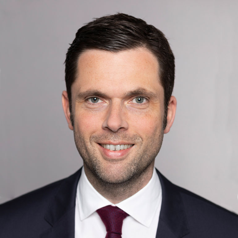 Ein lächelnder Mann mit kurzem, dunklem Haar trägt einen Anzug und eine Krawatte. Die Hintergrundfarbe ist hellgrau. Der Mann sieht direkt in die Kamera und vermittelt einen freundlichen Eindruck.