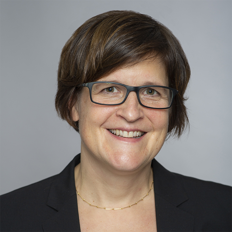 Porträt einer lächelnden Frau mit kurzen braunen Haaren und Brille, die einen schwarzen Blazer trägt. Der Hintergrund ist hellgrau, was den Fokus auf ihr freundliches Gesicht lenkt.