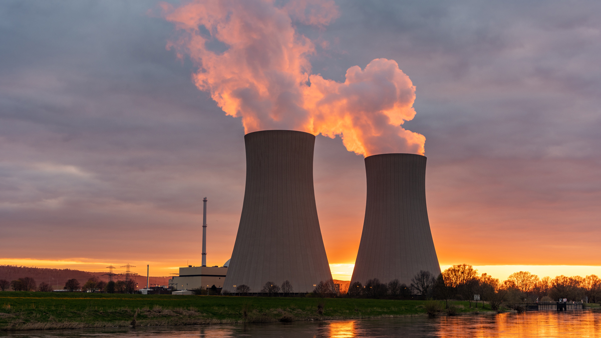 Zwei Kühltürme eines Kernkraftwerks stehen im Vordergrund, aus denen Rauch aufsteigt. Im Hintergrund ist ein Sonnenuntergang zu sehen, der den Himmel in warmen Farbnuancen leuchtet. Die Landschaft umfasst einen Fluss und teilweise bewaldete Ufer.