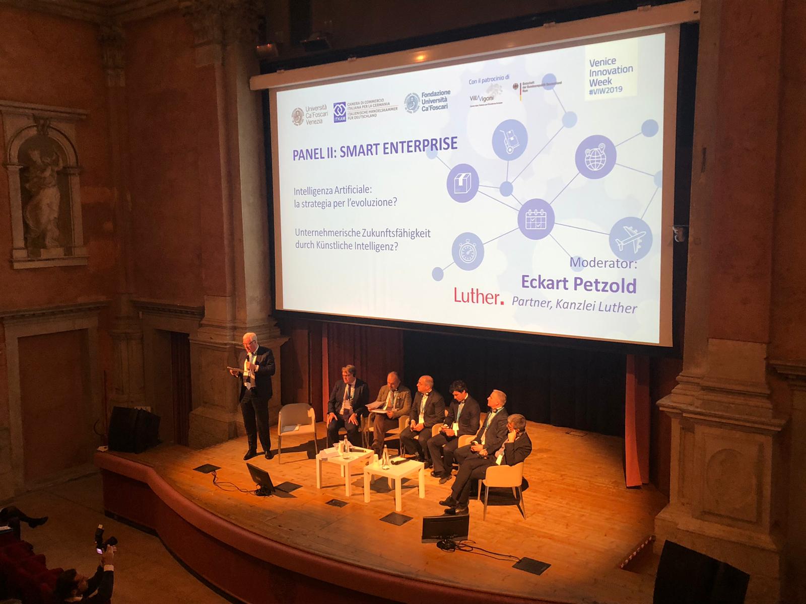 A panel discussion titled "Smart Enterprise" takes place on stage, featuring several speakers seated together. The moderator, Eckart Petzold, stands and addresses the audience. A large screen behind displays topics related to artificial intelligence and its impact on business evolution.