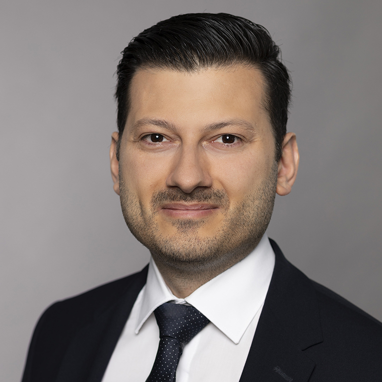 Ein Mann mit kurzen, dunklen Haaren trägt einen Anzug und eine Krawatte. Er steht vor einem neutralen, grauen Hintergrund und lächelt leicht in die Kamera. Sein Ausdruck ist freundlich und selbstbewusst.