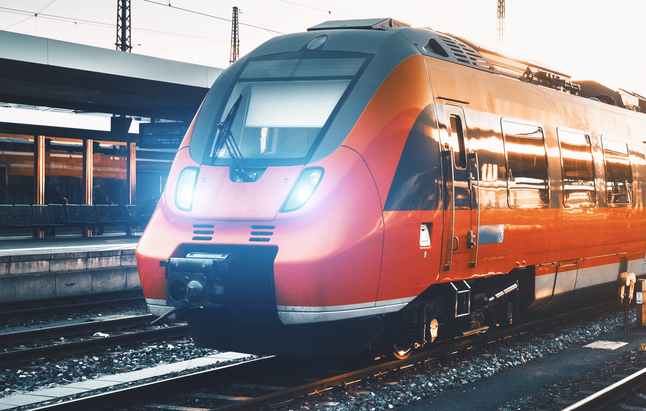Ein roter Zug steht auf einem Bahnhofsgleis. Die Front ist gut sichtbar, mit leuchtenden Scheinwerfern. Im Hintergrund sind Gleise und Teile des Bahnhofs zu erkennen. Die Stimmung ist durch das Tageslicht und die Umgebung geprägt.