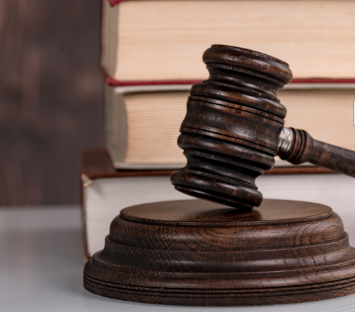 A wooden gavel is placed on a circular base, symbolizing justice and authority. Behind it are stacked law books, suggesting a legal context. The image conveys themes of law, courtroom proceedings, and the legal profession.