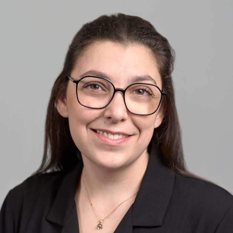 Porträt einer lächelnden jungen Frau mit langen, dunklen Haaren, die eine schwarze Bluse trägt und eine feine Kette um den Hals hat. Sie trägt eine modische Brille und hat einen freundlichen Ausdruck vor einem grauen Hintergrund.