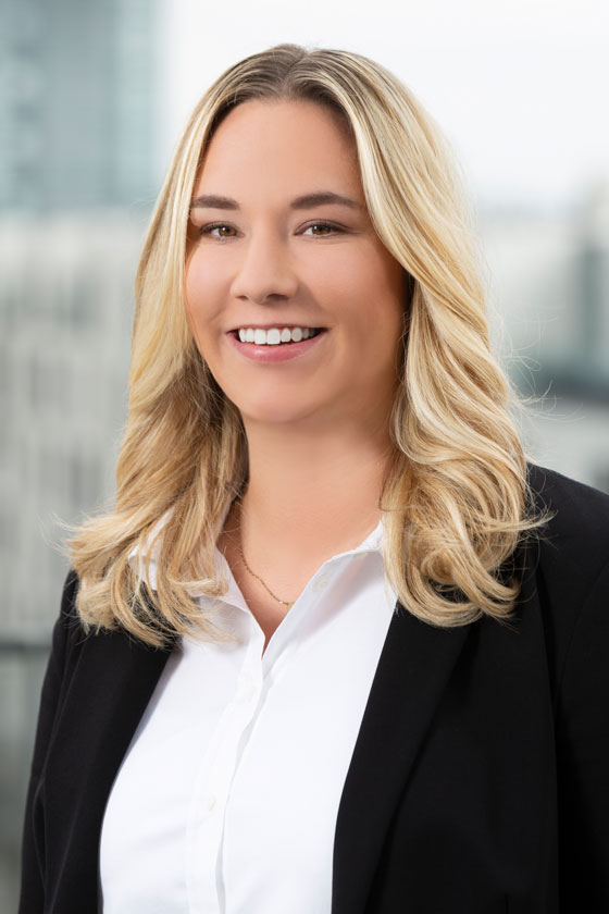 Ein Porträt einer lächelnden Person mit langen, blonden Haaren, die ein weißes Hemd und einen schwarzen Blazer trägt. Der Hintergrund zeigt unscharfe architektonische Elemente, was auf eine urbane Umgebung hinweist.