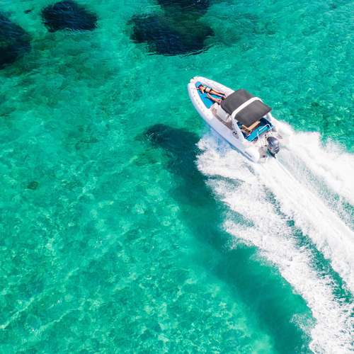 Ein Motorboot fährt über das klare, türkisfarbene Wasser, hinterlässt Gischt und Wellen. Unter dem Wasser sind dunklere Flecken sichtbar, die auf den Grund hinweisen.