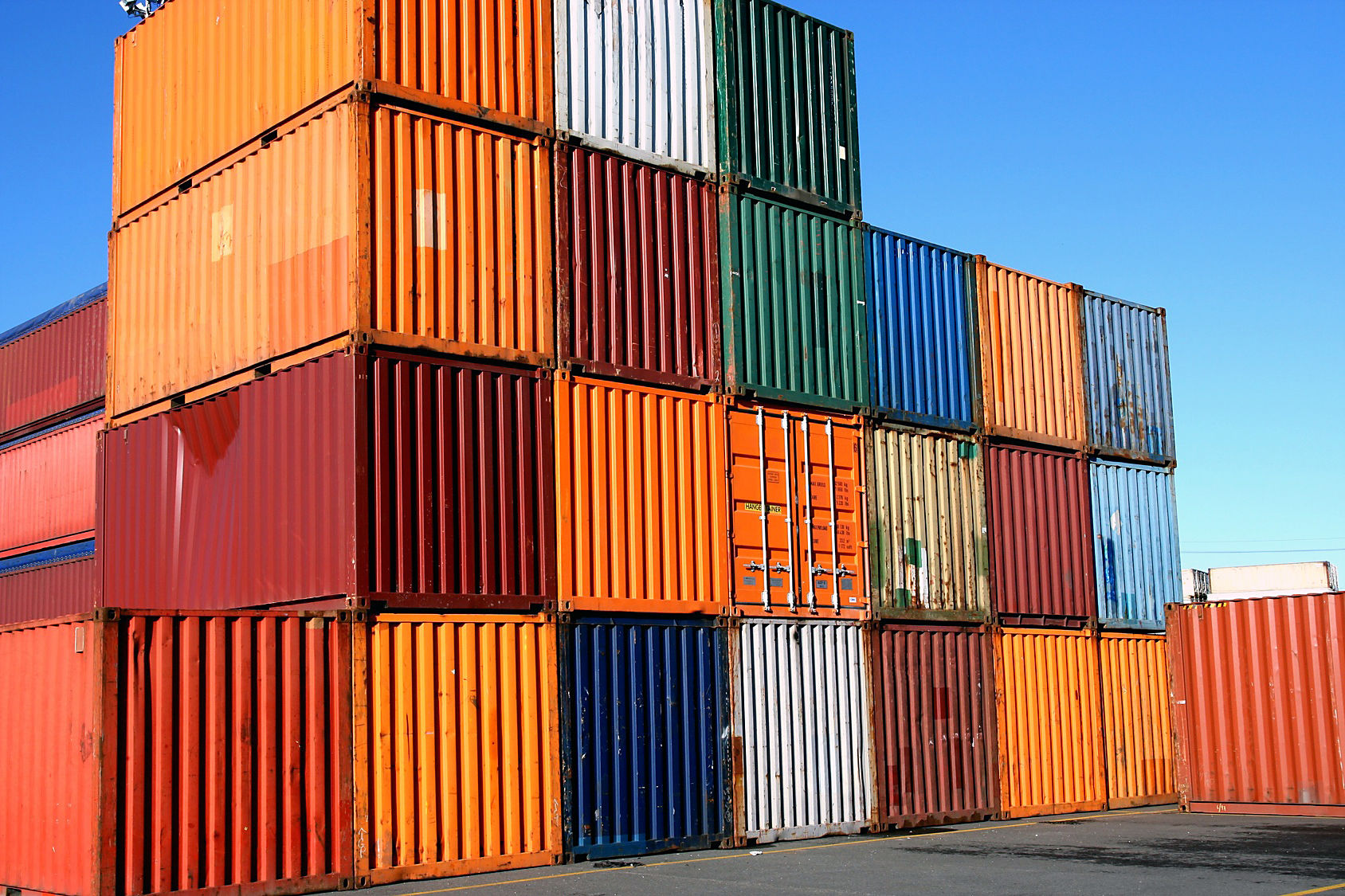 A stack of shipping containers in various colors, including orange, blue, green, red, and white, is arranged in a grid-like pattern against a clear blue sky. The containers vary in height and alignment, creating a textured and visually striking wall.