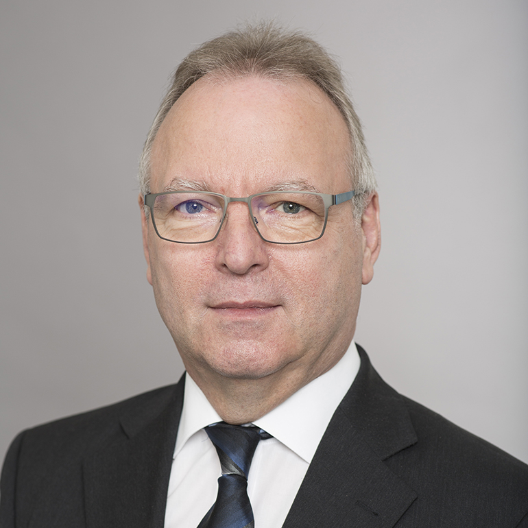 Ein Mann mit Brille und grauen Haaren trägt einen Anzug und eine Krawatte. Er blickt direkt in die Kamera vor einem neutralen Hintergrund. Der Ausdruck ist seriös und professionell.