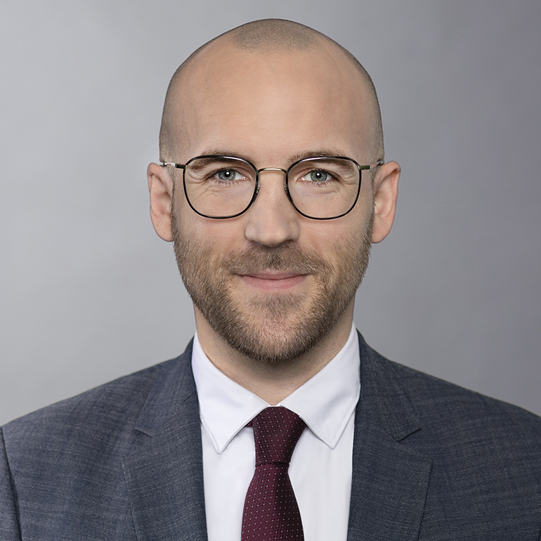 Ein lächelnder Mann mit Glatze und Brille trägt einen Anzug und eine Krawatte. Der Hintergrund ist neutral grau, was den Fokus auf sein freundliches Gesicht lenkt.