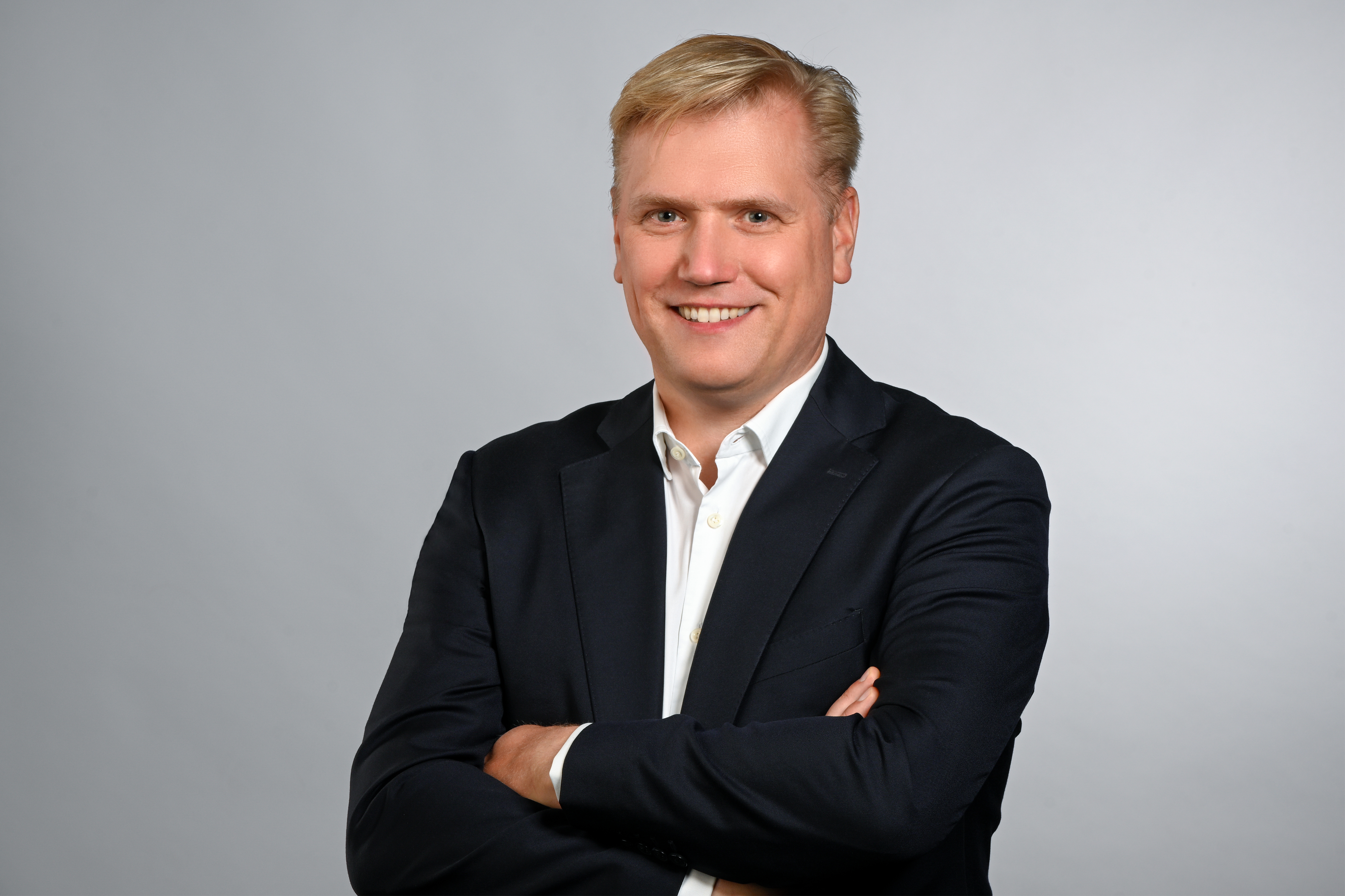 A professional man smiles confidently while standing with arms crossed, dressed in a dark suit over a white shirt. The background is a soft, light gray.