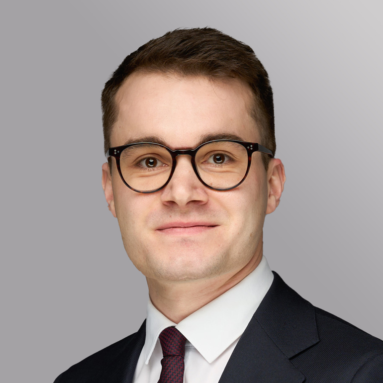 Ein junger Mann mit Brille lächelt in die Kamera. Er trägt ein Hemd und eine Krawatte, sowie einen Anzug. Der Hintergrund ist grau.