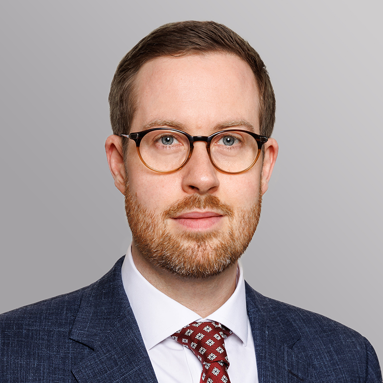 Porträt eines Mannes mit Brille und Bart. Er trägt einen Anzug und ein geblümtes Hemd. Der Hintergrund ist in sanften Grautönen gehalten. Der Mann schaut neutral in die Kamera.