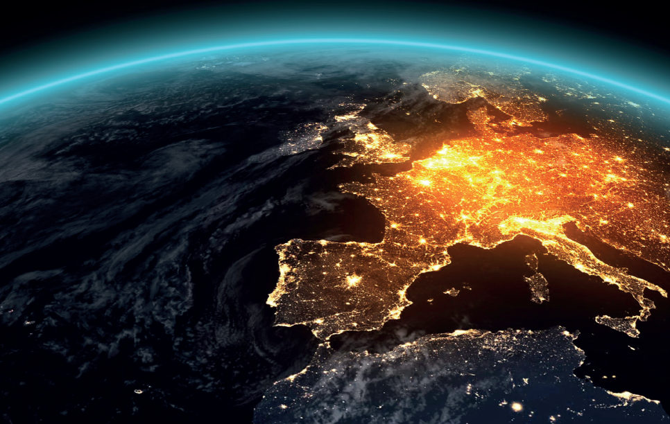 A view of Earth from space, showcasing a nighttime perspective of Europe illuminated by city lights. The scene highlights the contrast between the dark ocean and the vibrant, glowing regions of populated areas on the continent. The curvature of the planet is visible in the background.