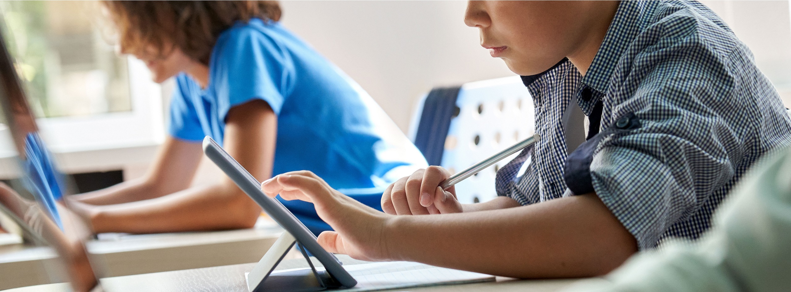 A close-up view of a child using a tablet for studying in a classroom setting. The child holds a stylus and interacts with the device, focused on their task, while another student is visible in the background, engaged in similar activities.