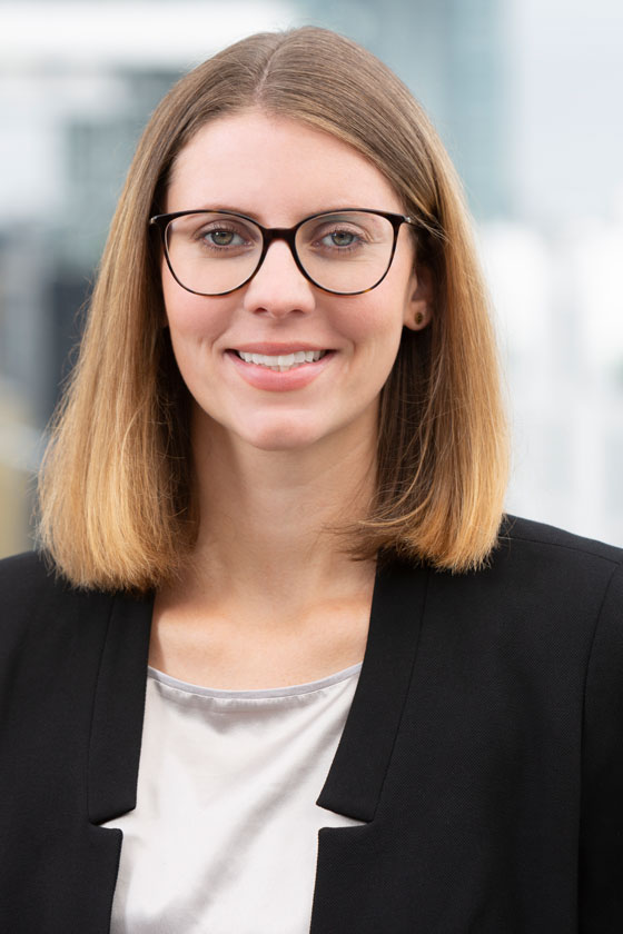 Portrait einer Frau mit schulterlangen, blonden Haaren und Brille. Sie trägt ein schwarzes Blazer und ein helles Oberteil. Der Hintergrund zeigt unscharfe Hochhäuser, was auf ein urbanes Umfeld hindeutet. Sie lächelt freundlich in die Kamera.