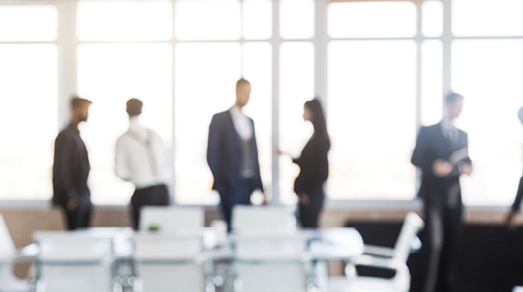 Zwei Gruppen von Personen in formeller Kleidung stehen in einem modernen Büroraum, wobei die Fenster im Hintergrund viel Licht hereinlassen. Einige Menschen unterhalten sich, während andere auf ihre mobilen Geräte schauen. Tische mit Stühlen sind im Vordergrund sichtbar.