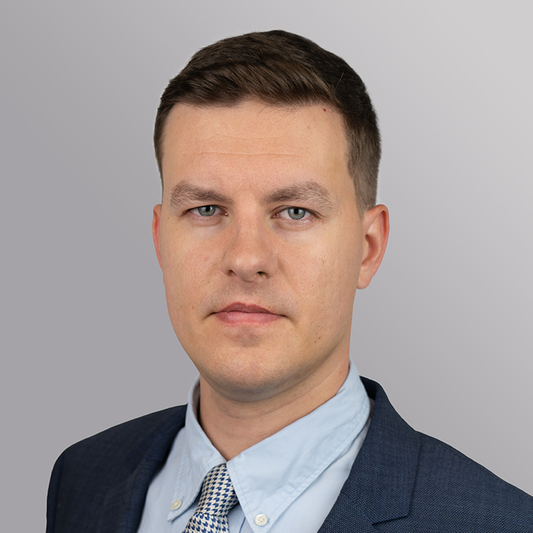 Ein junger Mann mit kurzem, braunem Haar trägt einen dunklen Anzug und ein helles Hemd mit Krawatte. Er blickt direkt in die Kamera und präsentiert einen selbstbewussten Ausdruck. Der Hintergrund ist grau gehalten.