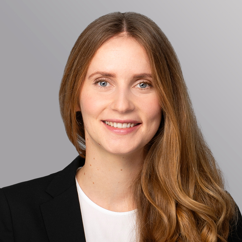 Portrait einer Frau mit langen, welligen Haaren, die freundlich lächelt. Sie trägt ein dunkles Jacket über einem weißen Oberteil. Der Hintergrund ist in sanften Grautönen gehalten.