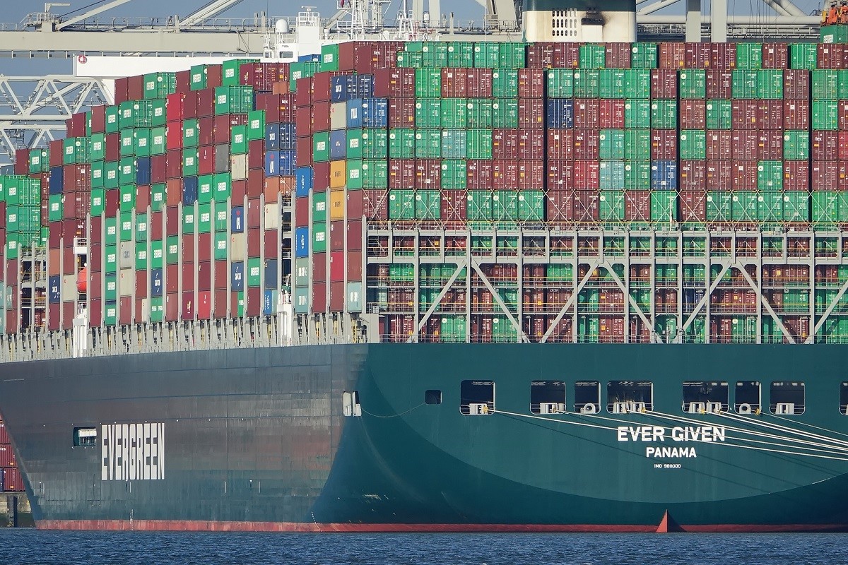 Ein großes Containerschiff mit der Aufschrift "EVER GIVEN" und einer Vielzahl an bunten Containern in grünen, roten und blauen Farben. Das Schiff ist an einer modernen Hafeneinrichtung angelegt, bei klarem Himmel und ruhigem Wasser.