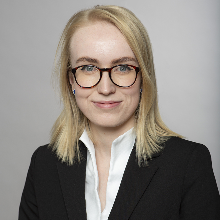 Porträt einer Person mit blonden Haaren, die eine schwarze Jacke und ein weißes Hemd trägt. Sie hat eine freundliche Miene und trägt eine Brille. Der Hintergrund ist neutral und hell.