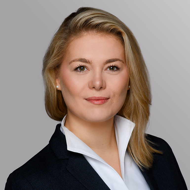 Ein Porträt einer Frau mit blonden Haaren, die in einem dunkelblauen Anzug und einem weißen Hemd gekleidet ist. Sie schaut direkt in die Kamera und hat einen freundlichen Gesichtsausdruck. Hintergrund in sanften Grautönen.