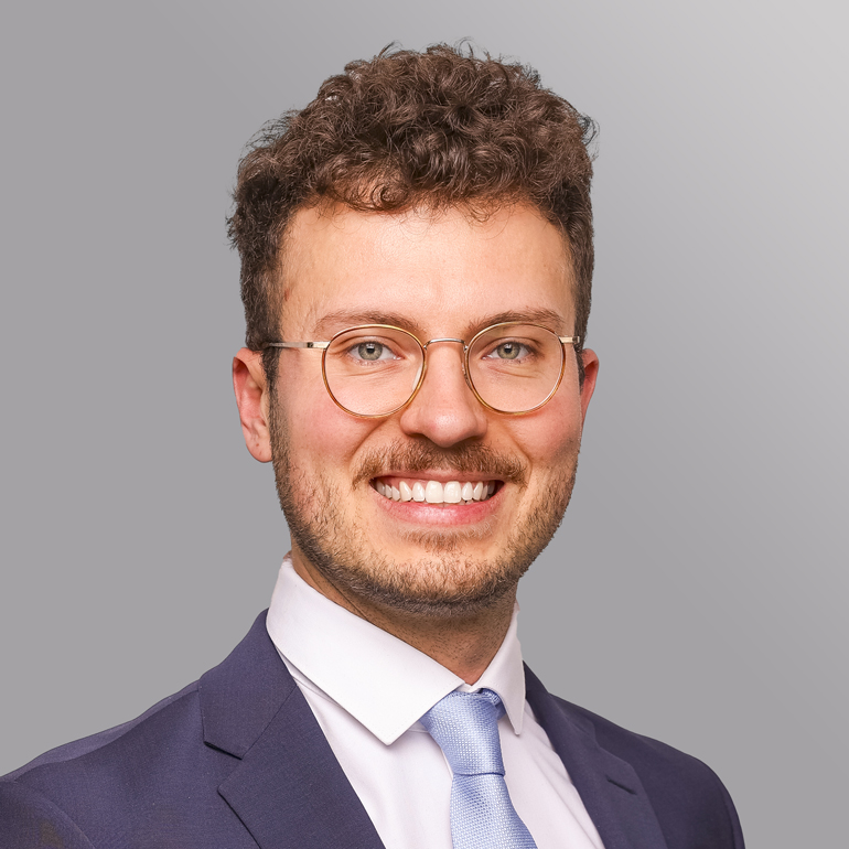 Porträt eines lächelnden Mannes mit lockigem, braunem Haar und Brille. Er trägt einen Anzug und eine Krawatte und hat einen freundlichen Ausdruck. Im Hintergrund ist eine graue, gleichmäßige Fläche.