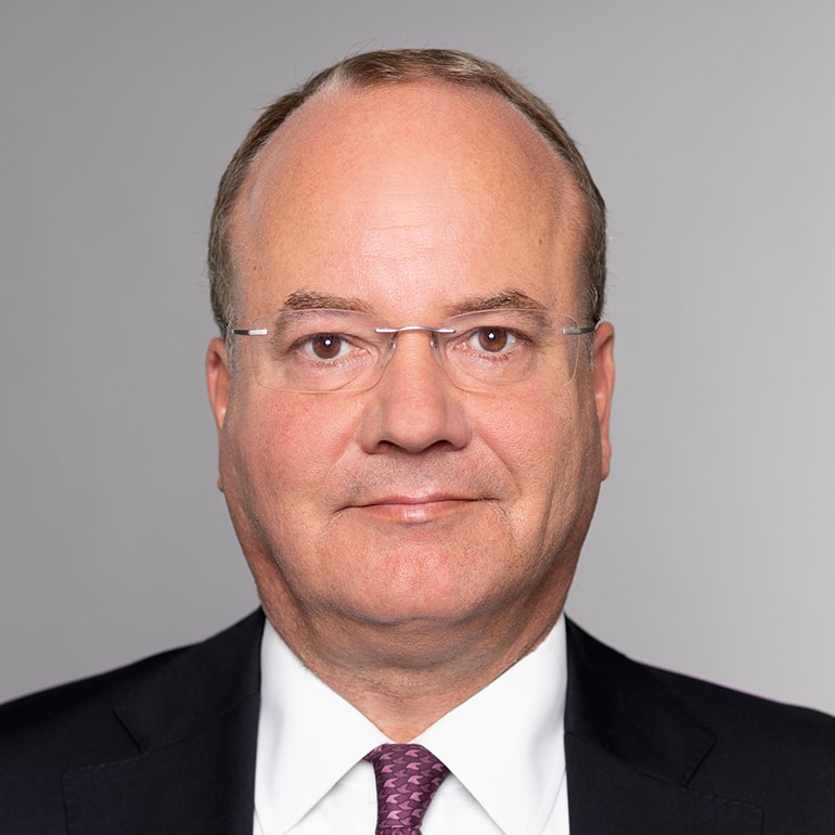 Portrait eines Mannes mit Brille und Anzug. Er hat glattes, hellbraunes Haar und einen neutralen Gesichtsausdruck. Der Hintergrund ist grau. Die gesamte Darstellung vermittelt einen professionellen Eindruck.