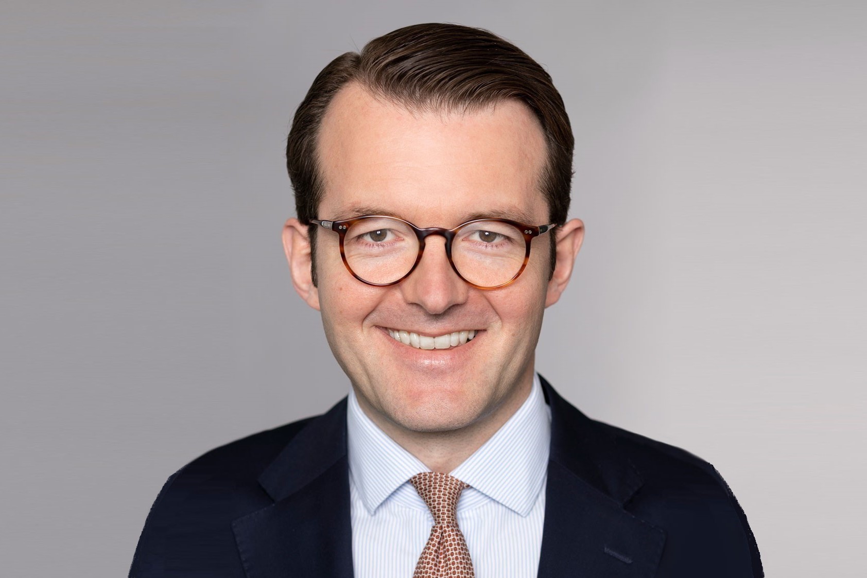 A smiling man with short, neatly styled hair and round, tortoiseshell glasses is wearing a dark suit, a light blue striped dress shirt, and a patterned tie. He is positioned against a neutral-colored background, exuding a professional and approachable demeanor.