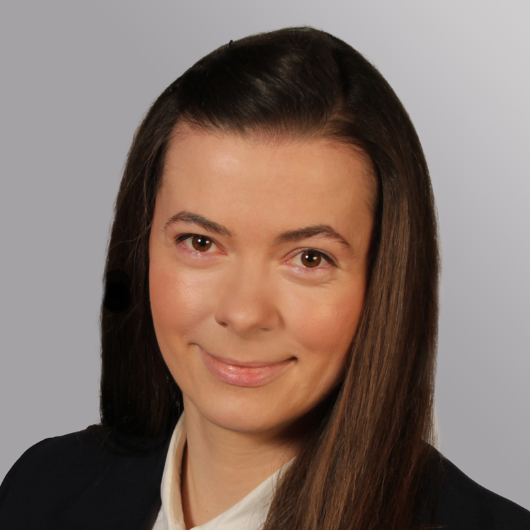 Portrait einer Frau mit langen, dunklen Haaren und einem freundlichen Lächeln. Sie trägt ein dunkelblaues Sakko und ein weißes Hemd. Der Hintergrund ist grau und weich verschwommen.