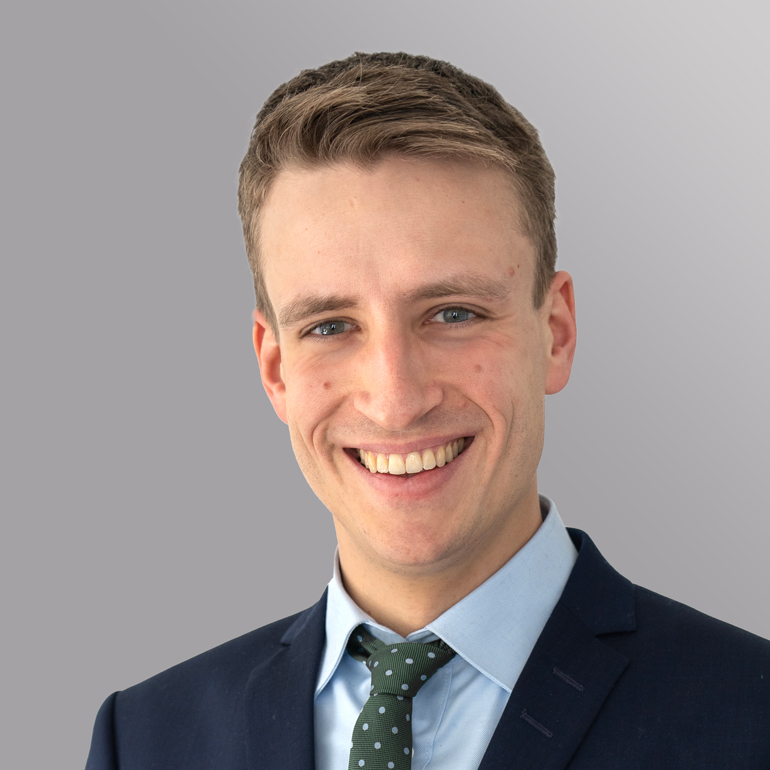 Ein junger Mann mit kurzem, hellbraunem Haar lächelt freundlich in die Kamera. Er trägt einen dunklen Anzug mit einem hellblauen Hemd und einer grün gepunkteten Krawatte. Der Hintergrund ist schlicht und grau.