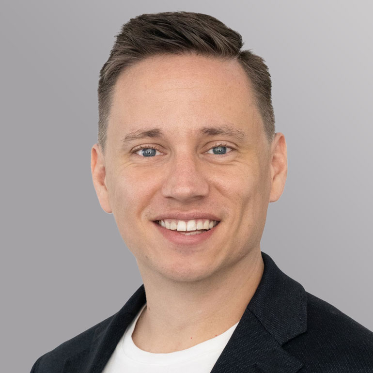 Porträt eines mannlichen Erwachsenen mit kurzen, blonden Haaren und blauem Hemd. Er lächelt freundlich in die Kamera, wobei ein neutraler, grauer Hintergrund zu sehen ist.