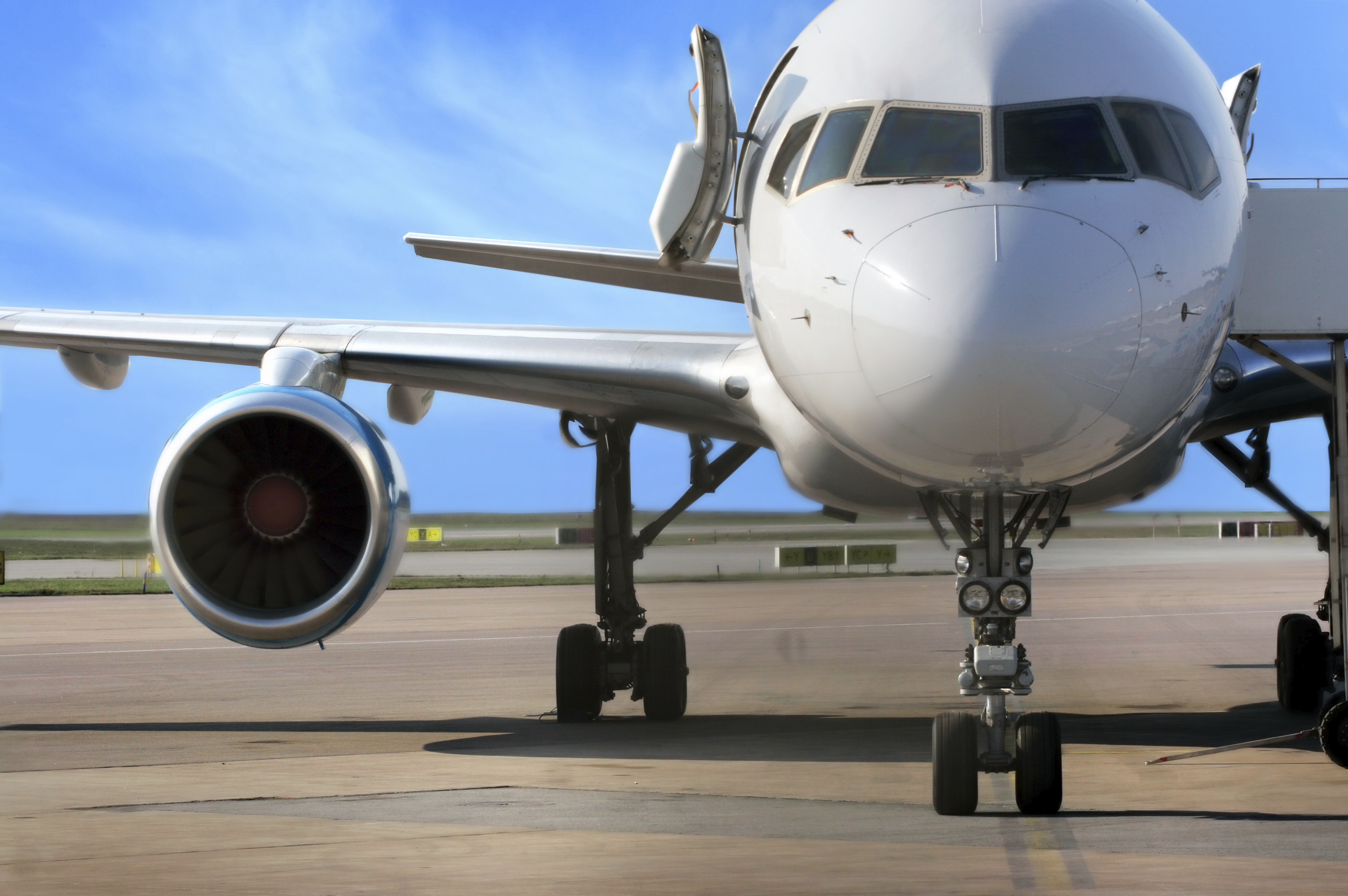 A commercial airplane is positioned on the runway, facing forward. The jet engine is visible under the wing, and the aircraft's front landing gear is on the ground, indicating readiness for takeoff. The background features a clear blue sky.