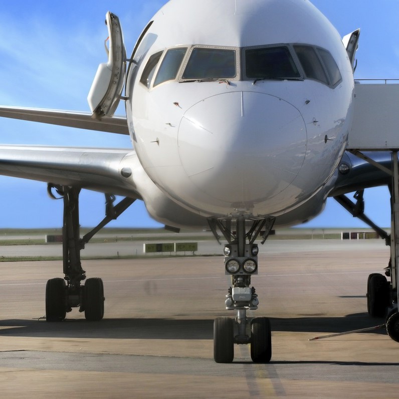 Ein Passagierflugzeug steht auf einer Landebahn. Die Vordertüren des Flugzeugs sind geöffnet, und man kann die Räder und den Schnauzenbereich deutlich sehen. Der Himmel im Hintergrund ist blau.
