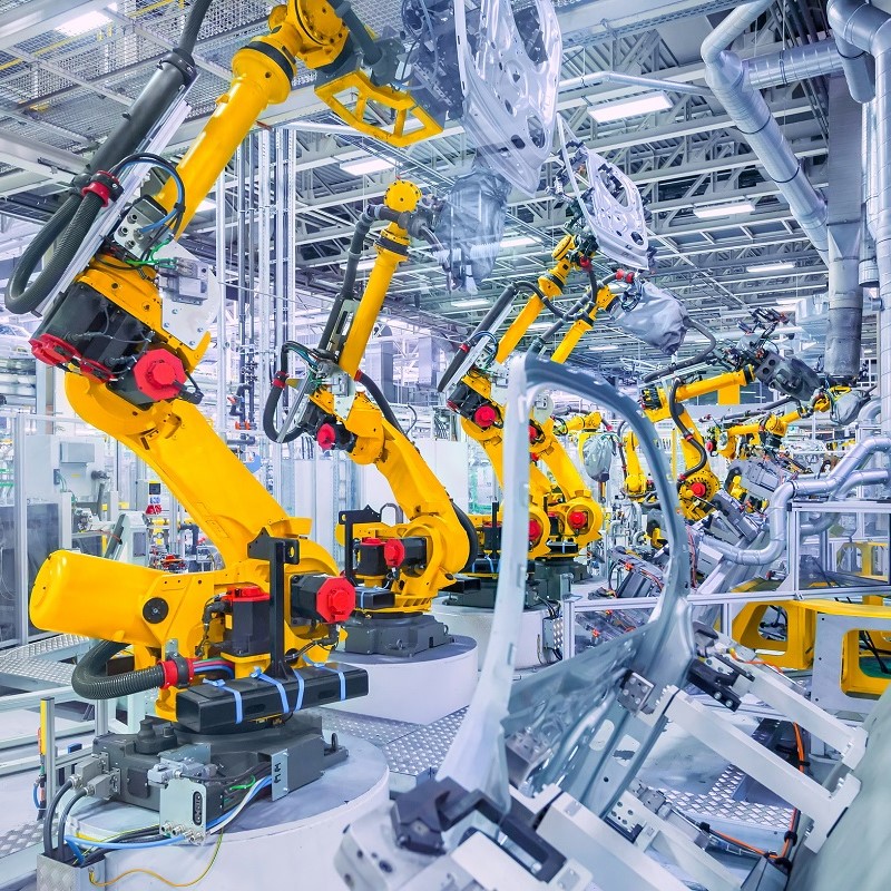 The image depicts a modern manufacturing facility filled with robotic arms. These yellow robotic arms are positioned on circular platforms, handling automotive parts in a high-tech assembly line. The environment is well-lit, showcasing a clean and organized industrial setting.