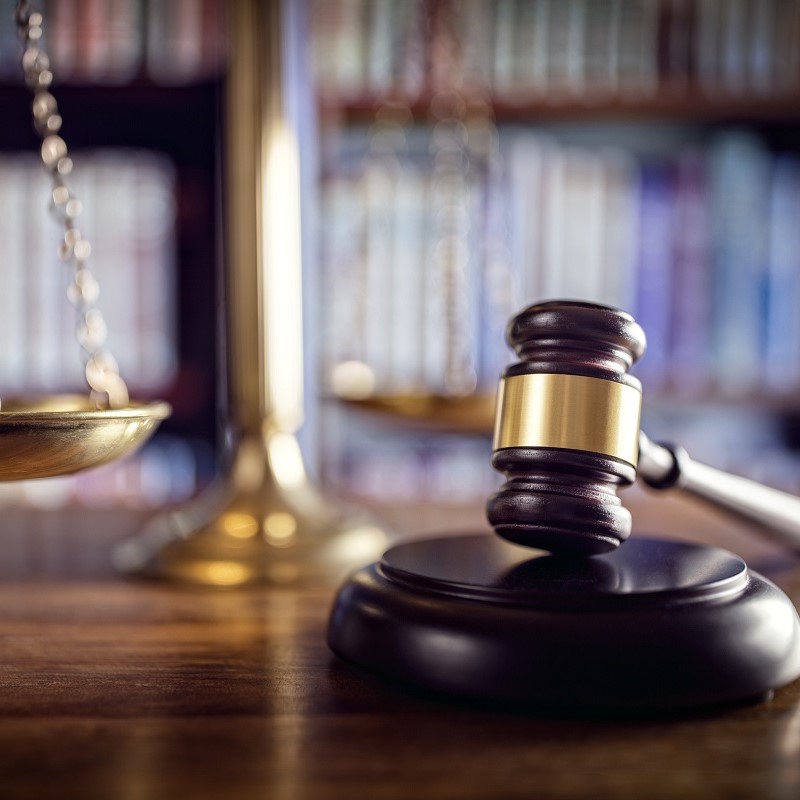 A wooden gavel rests on a round base, symbolizing justice and the legal system. In the background, a scale of justice is partially visible, highlighting the balance between rights and laws. The setting suggests a courtroom or legal environment.