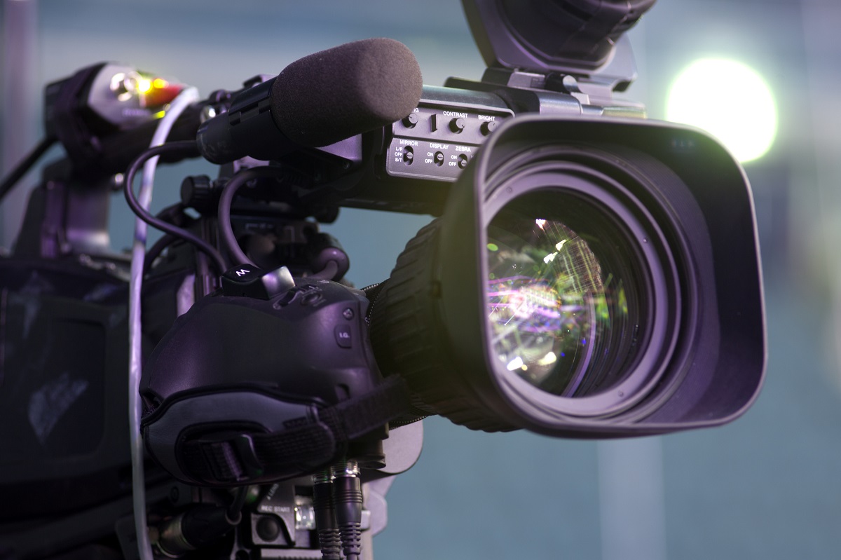A professional video camera is prominently displayed, featuring a large lens at the front and a microphone attached to the side. The camera has various buttons and controls visible, indicating its advanced functionality for recording audio and video. Soft lighting highlights its sleek design.