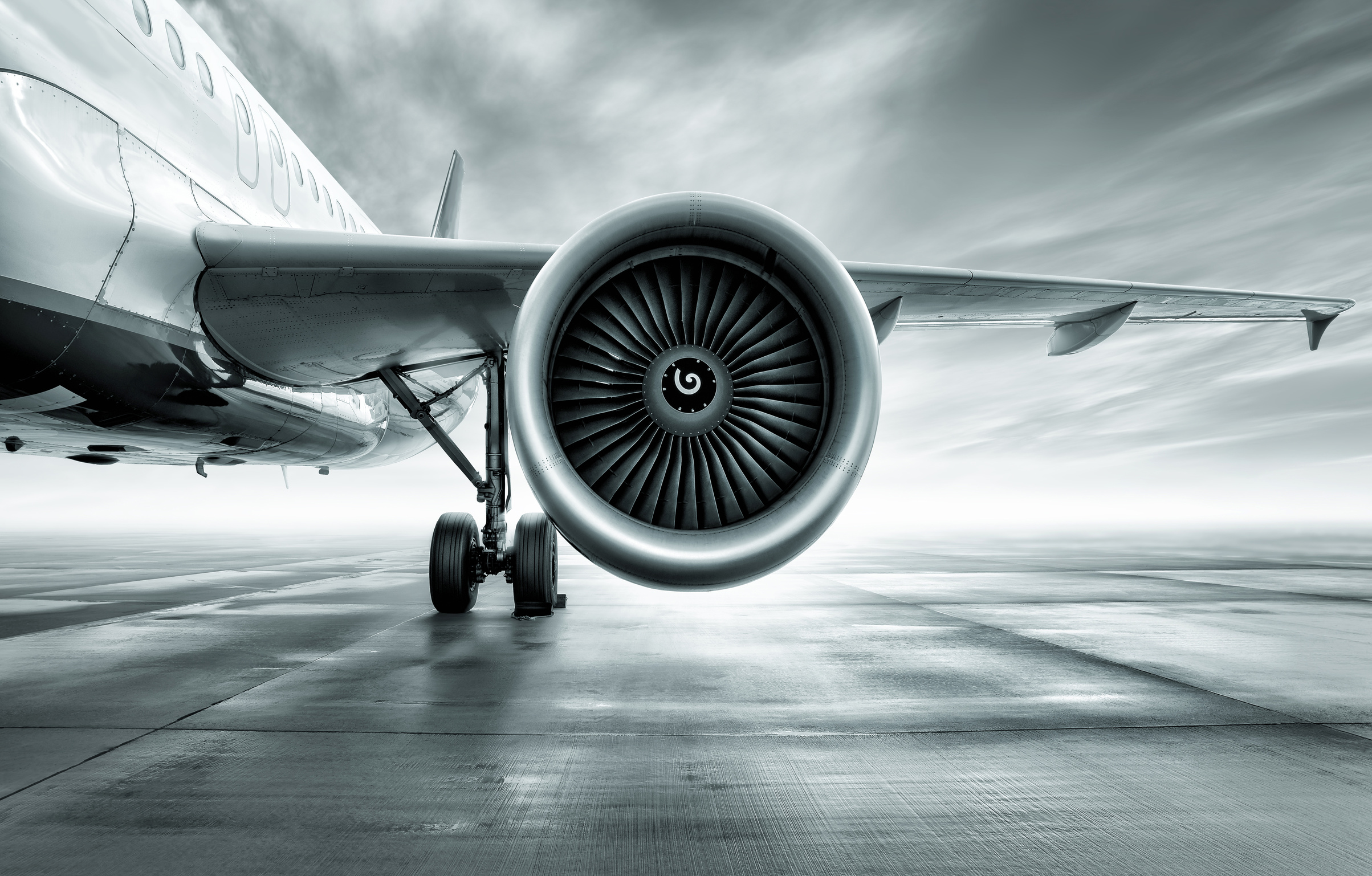 A close-up view of a commercial airplane's jet engine and wing. The image emphasizes the detailed structure of the engine and the aircraft's fuselage in a monochromatic color scheme against a blurred airport background.