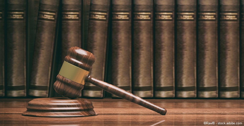 Ein Richterhammer liegt auf einer Holzplatte, daneben steht ein Zettel. Im Hintergrund sind mehrere große Bücher sichtbar, die eine rechtliche Themen behandeln. Die Szene vermittelt einen juristischen Kontext.