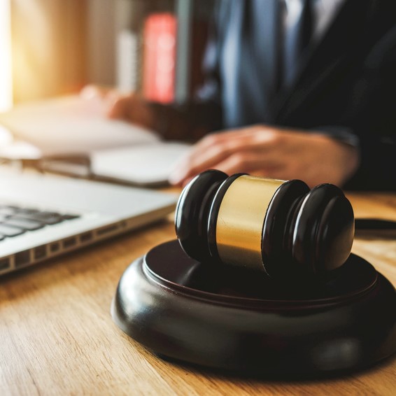 A wooden gavel with a gold band rests on a table in a legal office. In the background, a person in business attire writes in a notebook near a laptop, symbolizing legal work and decision-making.