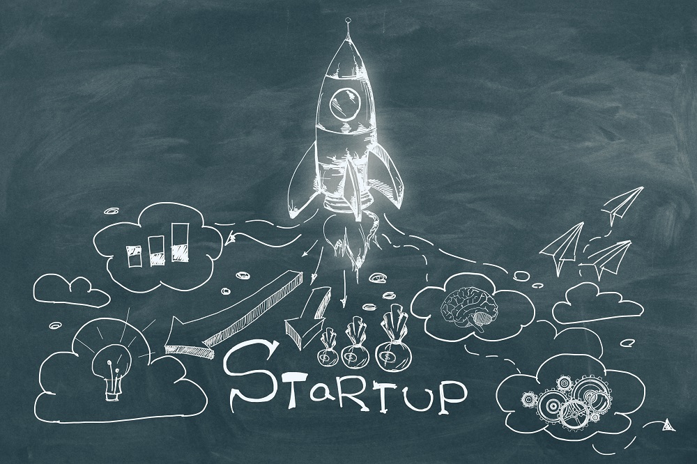 A chalkboard illustration depicts a rocket taking off labeled "STARTUP." Surrounding clouds contain symbols representing ideas, growth, and innovation, including light bulbs, graphs, and gears. The image conveys themes of entrepreneurship and progress.