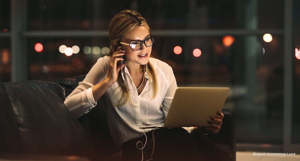 A woman with long hair and glasses sits in a darkened room, speaking on a phone while working on a laptop. She appears focused and engaged, with earphones connected. Soft, blurred lights can be seen in the background, suggesting an evening setting.