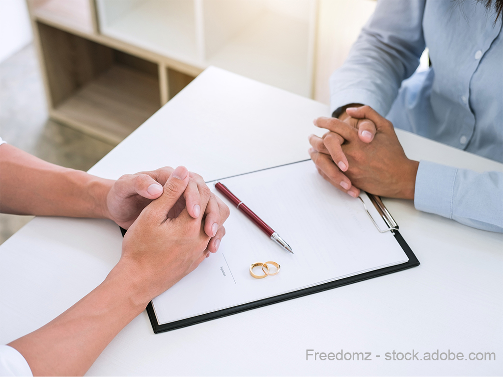 Zwei Personen sitzen sich an einem Tisch gegenüber. Ihre Hände sind gefaltet und ruhen auf einem Dokument. Auf dem Tisch liegt ein Stift und zwei Eheringe neben dem Dokument. Die Szene vermittelt eine angesannte Atmosphäre, möglicherweise während eines Gespräches über eine Trennung oder eine rechtliche Angelegenheit.