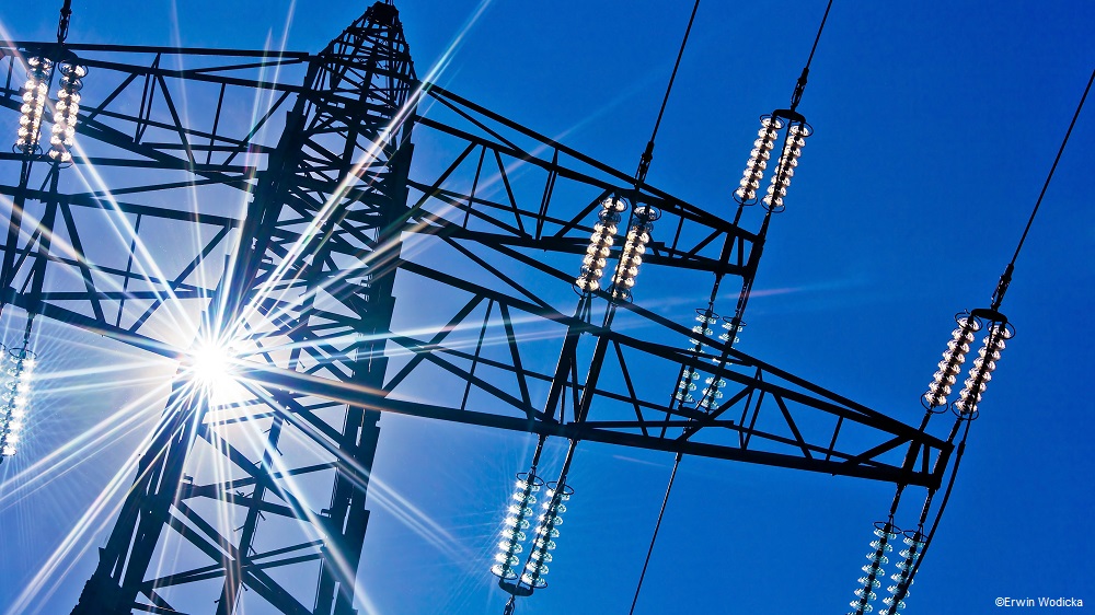 Eine Stromleitung aus Metall steht vor einem blauen Himmel. Sonnenstrahlen reflektieren auf den Lichtquellen, die an der Leitung montiert sind, und erzeugen einen strahlenden Effekt. Die hohe Struktur deutet auf elektrische Energieversorgung hin.
