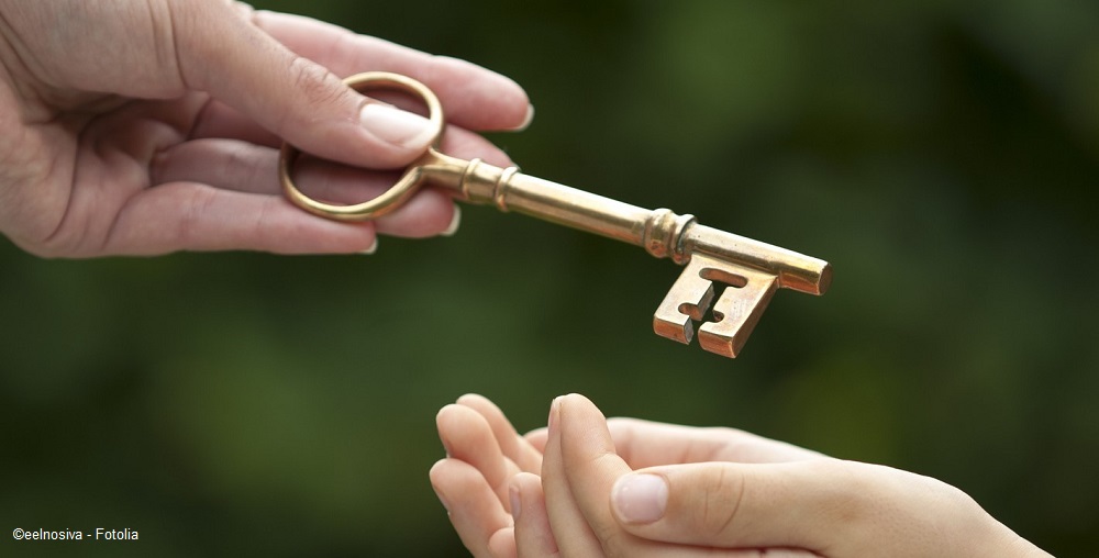 Eine Hand überreicht einen großen goldfarbenen Schlüssel an eine andere Hand. Der Schlüssel hat einen alten, dekorativen Stil und symbolisiert möglicherweise den Zugang oder eine wichtige Übergabe.