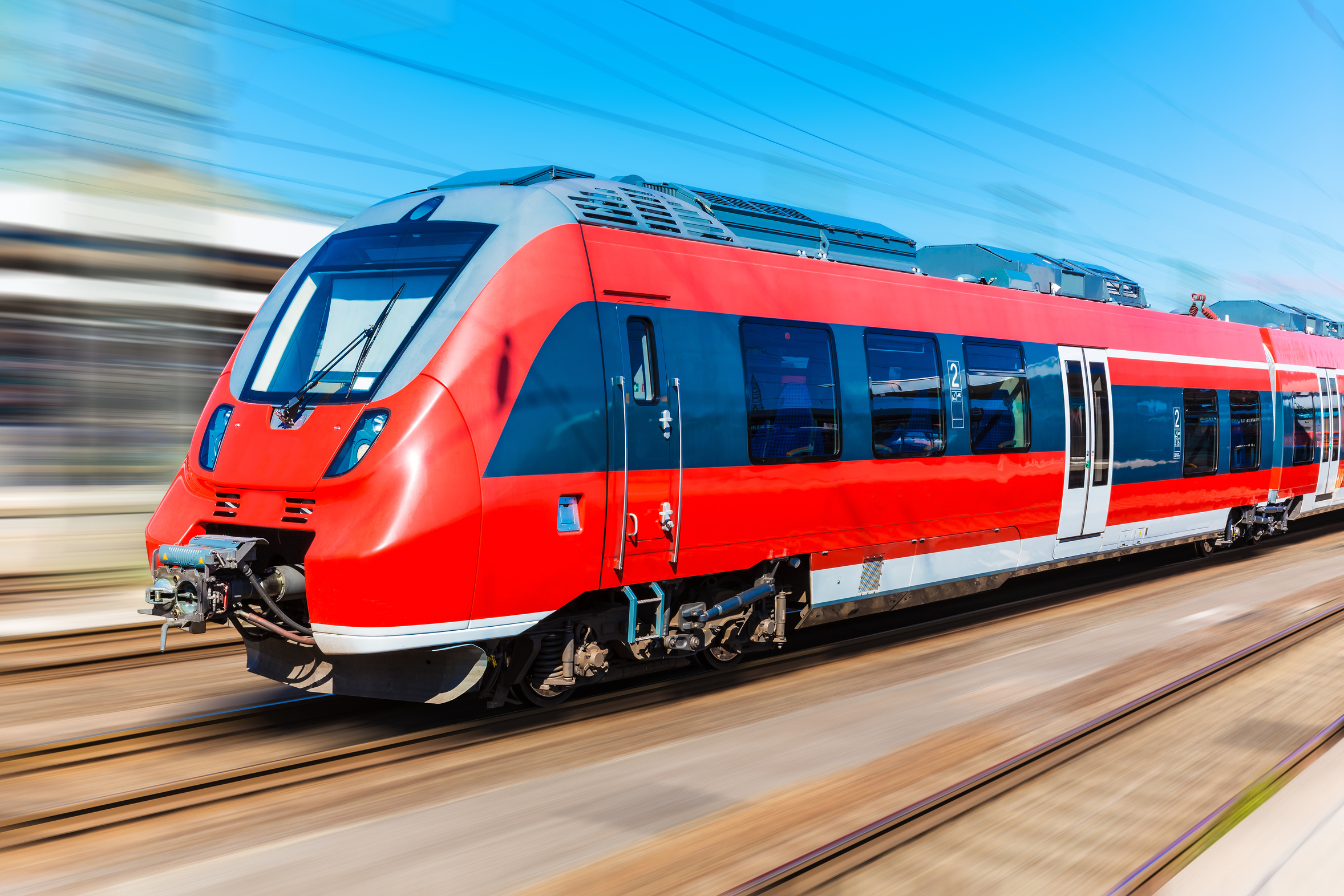 A red and gray passenger train is moving swiftly along the tracks, set against a bright blue sky. The image captures the train's streamlined design, with windows and doors visible, emphasizing its modern look and speed.