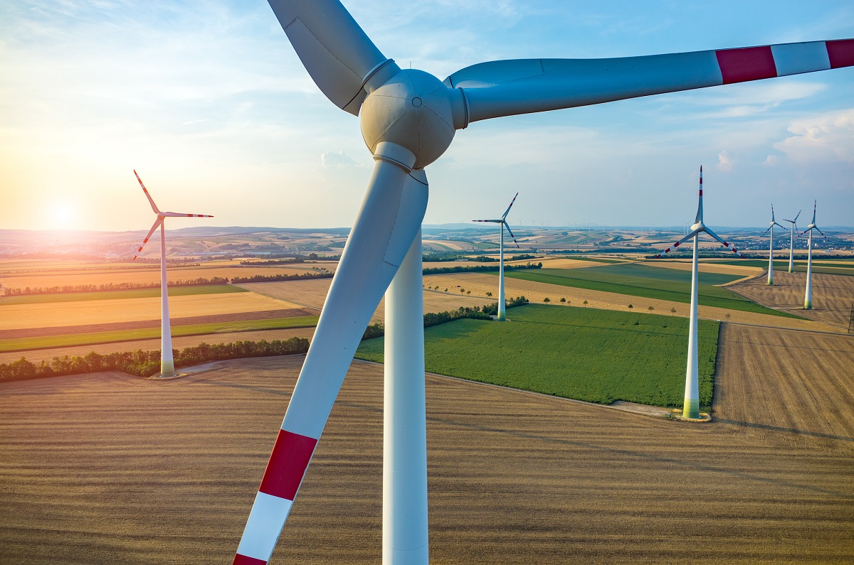 Aerial view of several wind turbines in a countryside landscape, with fields of green and brown stretching into the distance. The sun is setting, casting a warm glow over the scene, highlighting the alternative energy source's role in sustainable development.